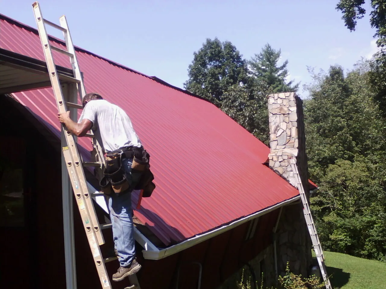Roofer on a ladder beside a red metal roof and stone chimney — Soffit & Fascia Repair by Roofing Brothers in Gray Court